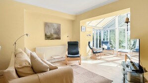 Een woonkamer met een bank, stoelen, een radiator, een schilderij en een glazen deur naar buiten.