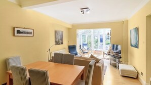 Een ruime kamer met een eettafel, stoelen, een bank en een televisie. Er zijn grote ramen en uitzicht op de omgeving.