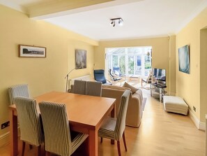 Ein geräumiger Raum mit Esstisch, Stühlen, einer Couch und einem Fernseher. Große Fenster bieten einen Blick ins Freie.