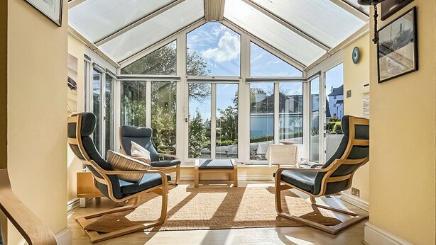 Een oranjerie met glazen dak, houten vloer en twee lounge fauteuils met kussens.