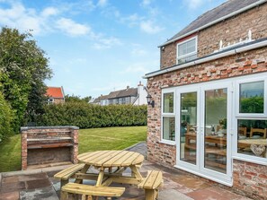 A brick house with a patio, a wooden table, and a barbecue area. - Old Master's Cottage (Leven)