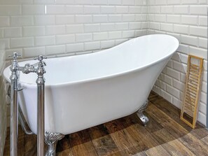 A white freestanding bathtub with ornate metal fixtures, a wooden floor, and a white tiled wall. - Old Master's Cottage (Leven)