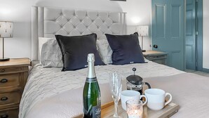 A neatly made bed with a tufted headboard, two dark pillows, a champagne bottle, a wine glass, and a tray with mugs and a coffee pot. - Old Master's Cottage (Leven)