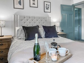 A neatly made bed with a tufted headboard, two dark pillows, a champagne bottle, a wine glass, and a tray with mugs and a coffee pot. - Old Master's Cottage (Leven)