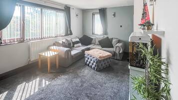 A living room with a sofa, ottoman, and a small table. There is a radiator, a window with curtains, and a plant.