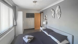 Ein Schlafzimmer mit einem Bett, einem Nachttisch mit Lampe, einem an der Wand befestigten Fernseher und dekorativer Wandgestaltung.