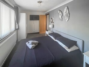 Ein Schlafzimmer mit einem Bett, einem Nachttisch mit Lampe, einem an der Wand befestigten Fernseher und dekorativer Wandgestaltung.