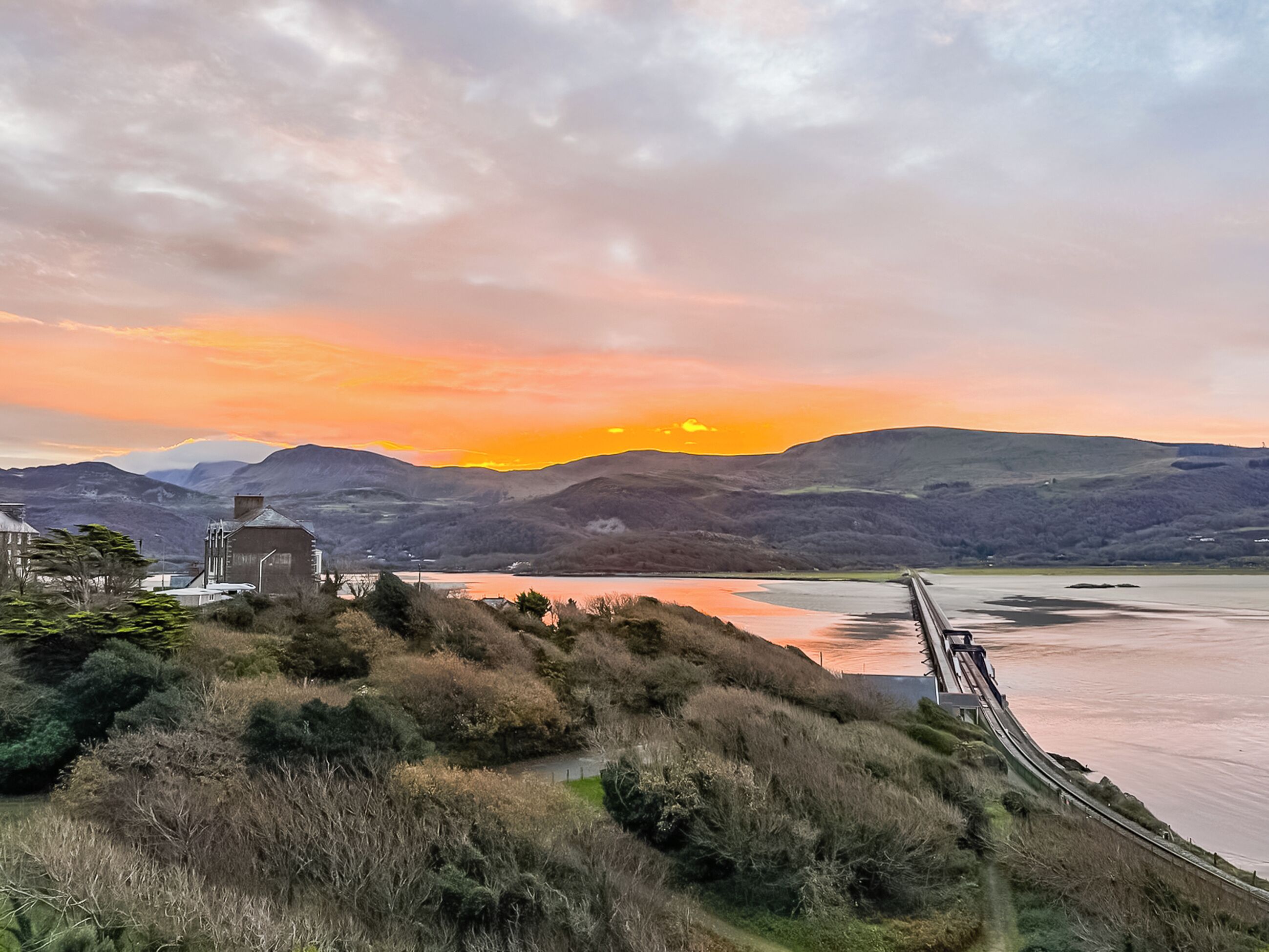 Eine malerische Landschaft mit einer Brücke, Hügeln und einem Gewässer bei Sonnenuntergang.