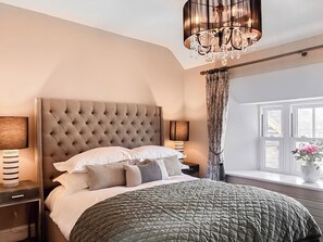 Ein Schlafzimmer mit einer gepolsterten Kopfteil, einem großen Bett mit vielen Kissen, Nachttischlampen, einem Fenster mit Vorhängen und einer Kristallleuchtre.