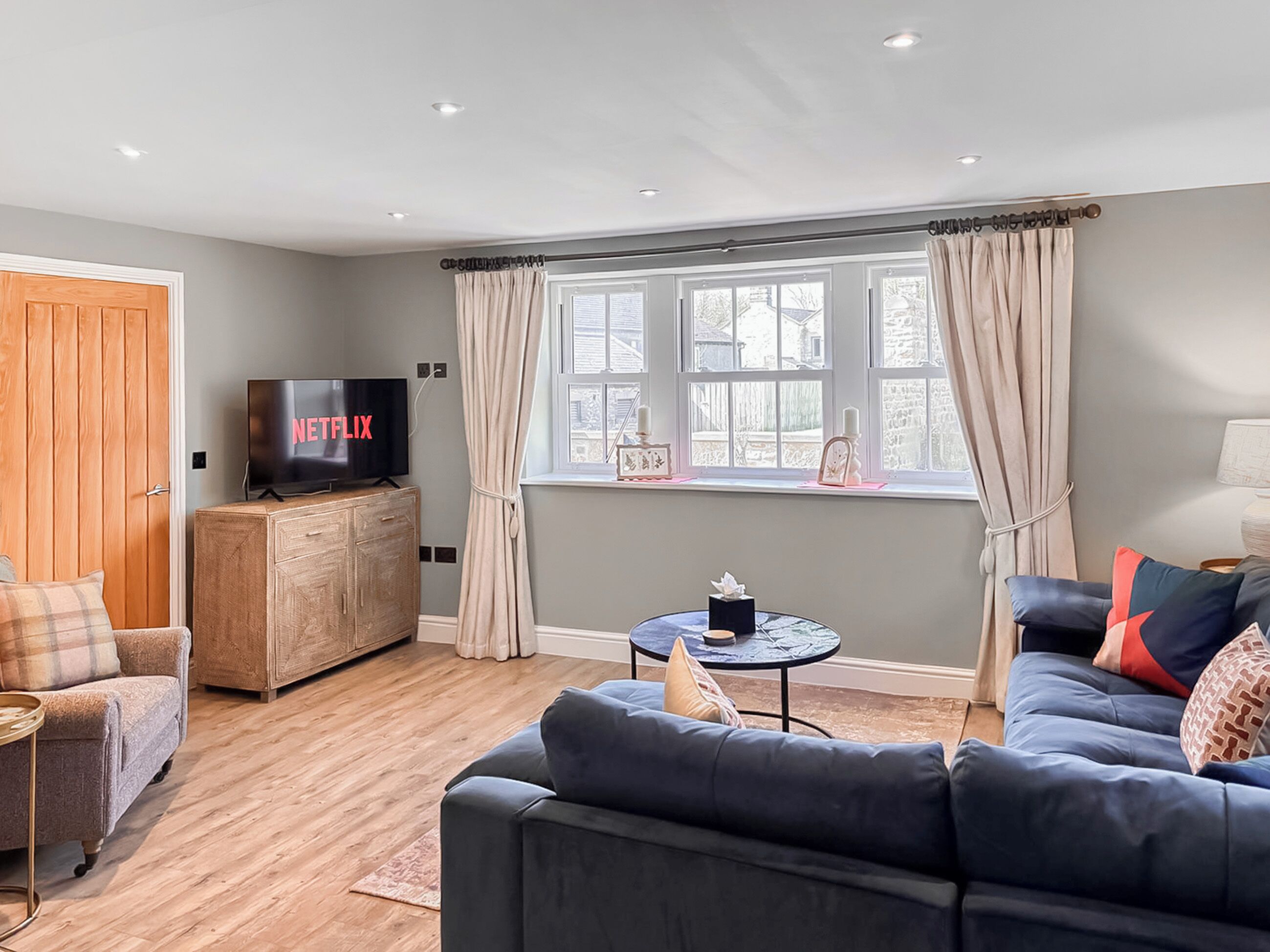 Una sala de estar con un sofá, un televisor sobre un mueble de madera, una pequeña mesa redonda y una ventana con cortinas.
