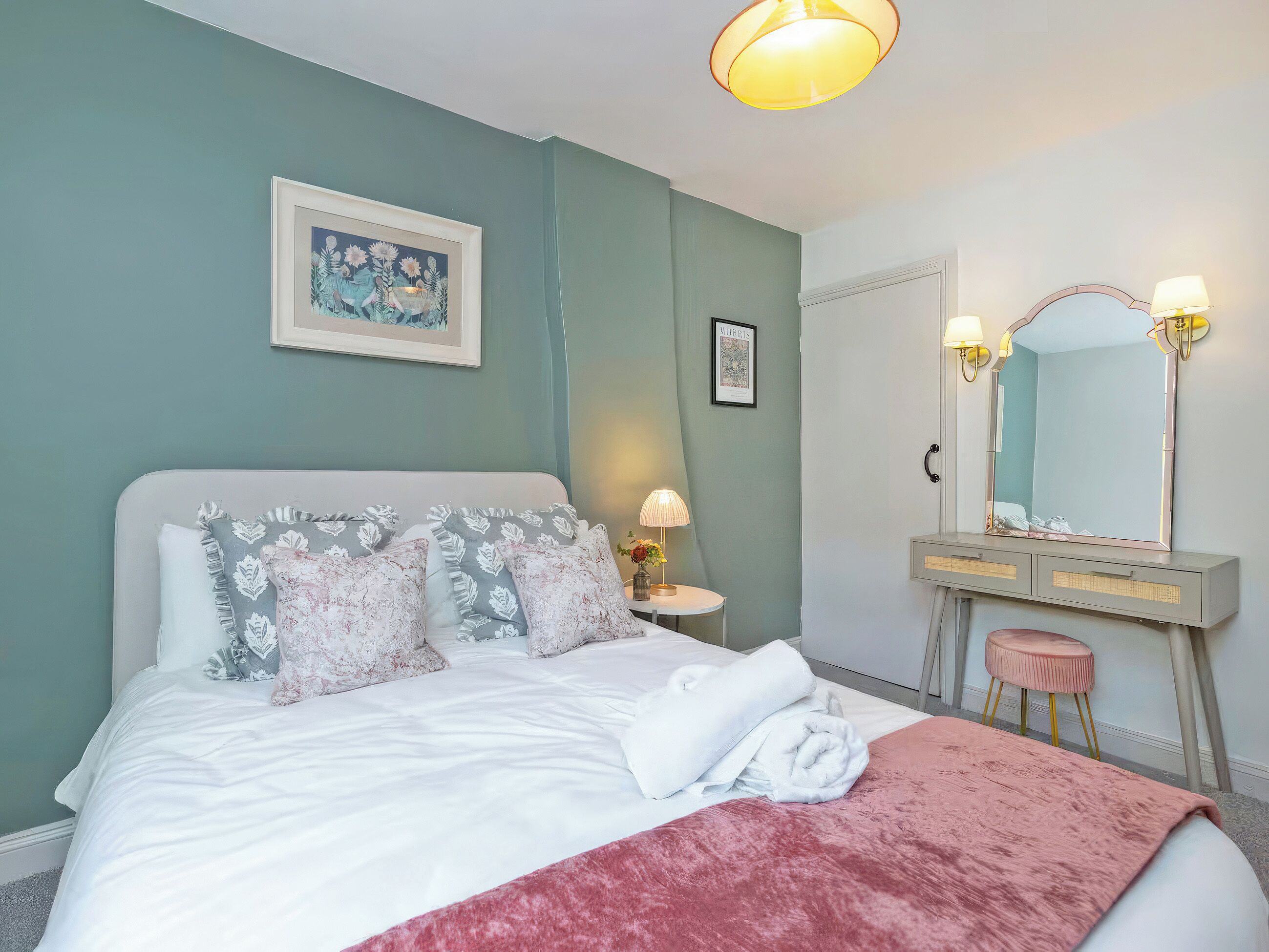 A neatly made bed with pillows, a bedside table with a lamp, a mirror with lights, and a framed picture on the wall.