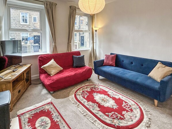 Une pièce avec deux canapés, une table d’appoint en bois, un tapis rouge et bleu, et une fenêtre avec des rideaux.