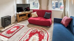 A living room with a red sofa, blue sofa, wooden entertainment unit, TV, and two rugs. - A Cosy two bedroom cottage, perfectly located in Bamford. (Bamford)