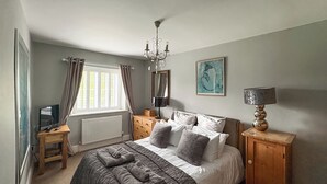 Un dormitorio con cama, una mesita de noche, una lámpara, un aparador, una ventana con cortinas y un cuadro en la pared.