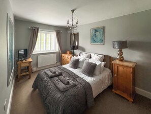 A bedroom with a bed, a bedside table, a lamp, a dresser, a window with curtains, and a painting on the wall. - Lower Park Farmhouse (Lyme Regis)