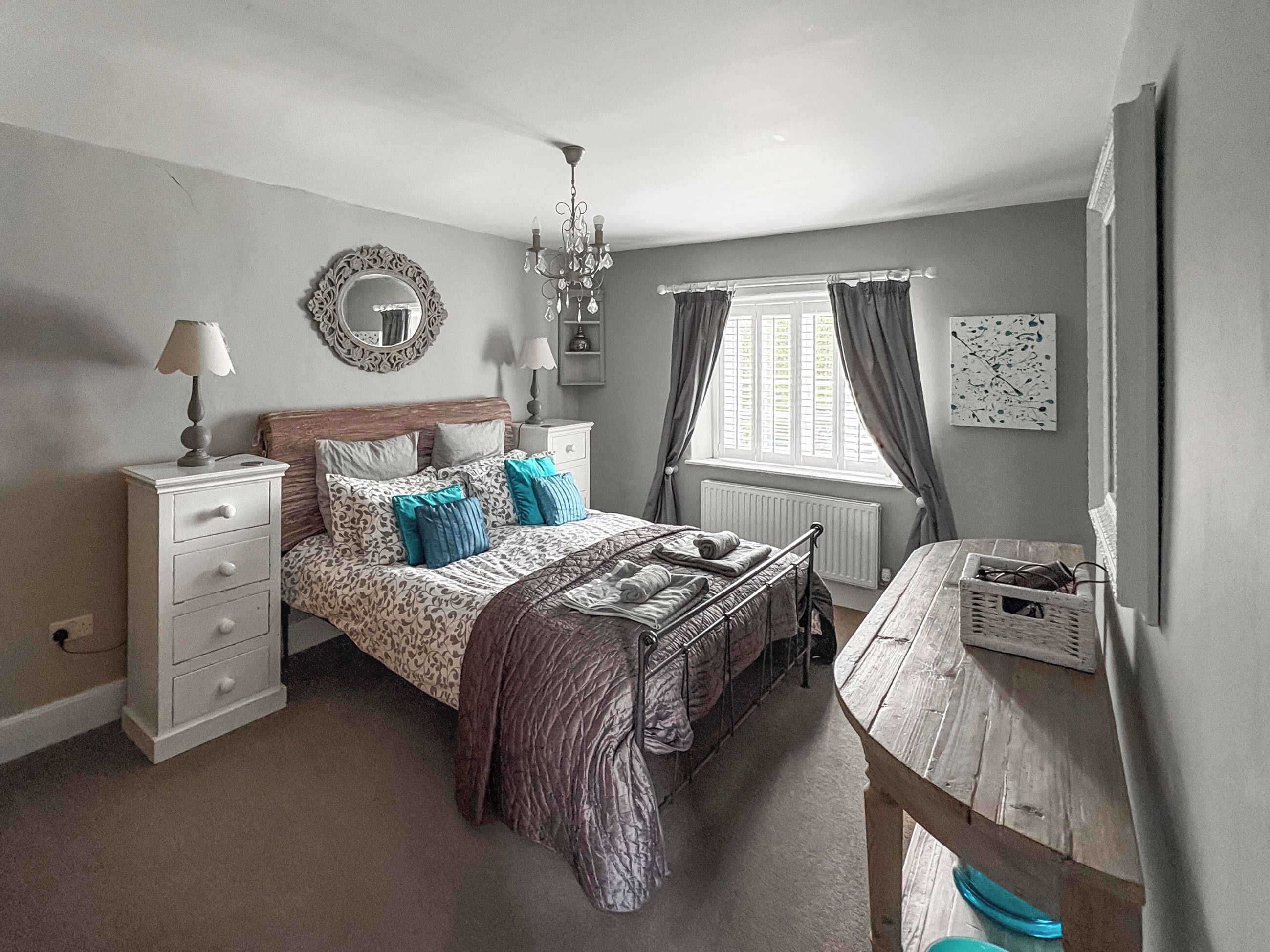 Une chambre à coucher comprenant un lit, des tables de chevet, une commode, un lustre, une fenêtre avec des rideaux et un miroir fixé au mur.