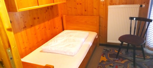 Un lit simple avec un drap blanc, une tête de lit en bois, une chaise avec un coussin et un radiateur au mur.