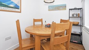 Een eetgedeelte met een ronde houten tafel en stoelen, een hanglamp en een plank met een radio en een kom.