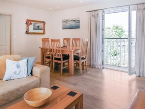 Ein Wohnzimmer mit einer Couch, einem Holzesstisch mit Stühlen, einem Balkon mit Vorhängen und zwei Bildern an der Wand.