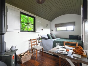 A cozy room with a bed, a table set for breakfast, a chair, a window with a view of greenery, and a wood-burning stove. - Bugail Hut - UK51876 (Cardiff)