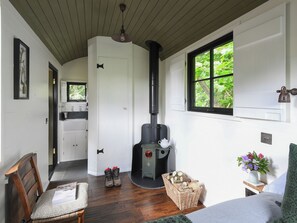 A room with a fireplace, a wooden chair, a bed, a window with a view of greenery, and a kitchen area. - Bugail Hut - UK51876 (Cardiff)