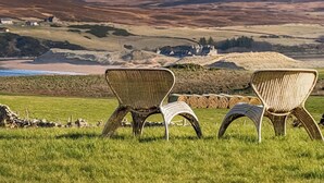 Drie rieten stoelen in een grasveld met uitzicht op glooiende heuvels en een gebouw in de verte.