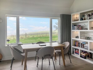 A dining area with a table and chairs, a bookshelf with books and decorative items, and a view of a green field through large windows.