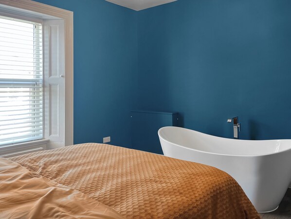 A room with a bed, a white bathtub, and a window with blinds.
