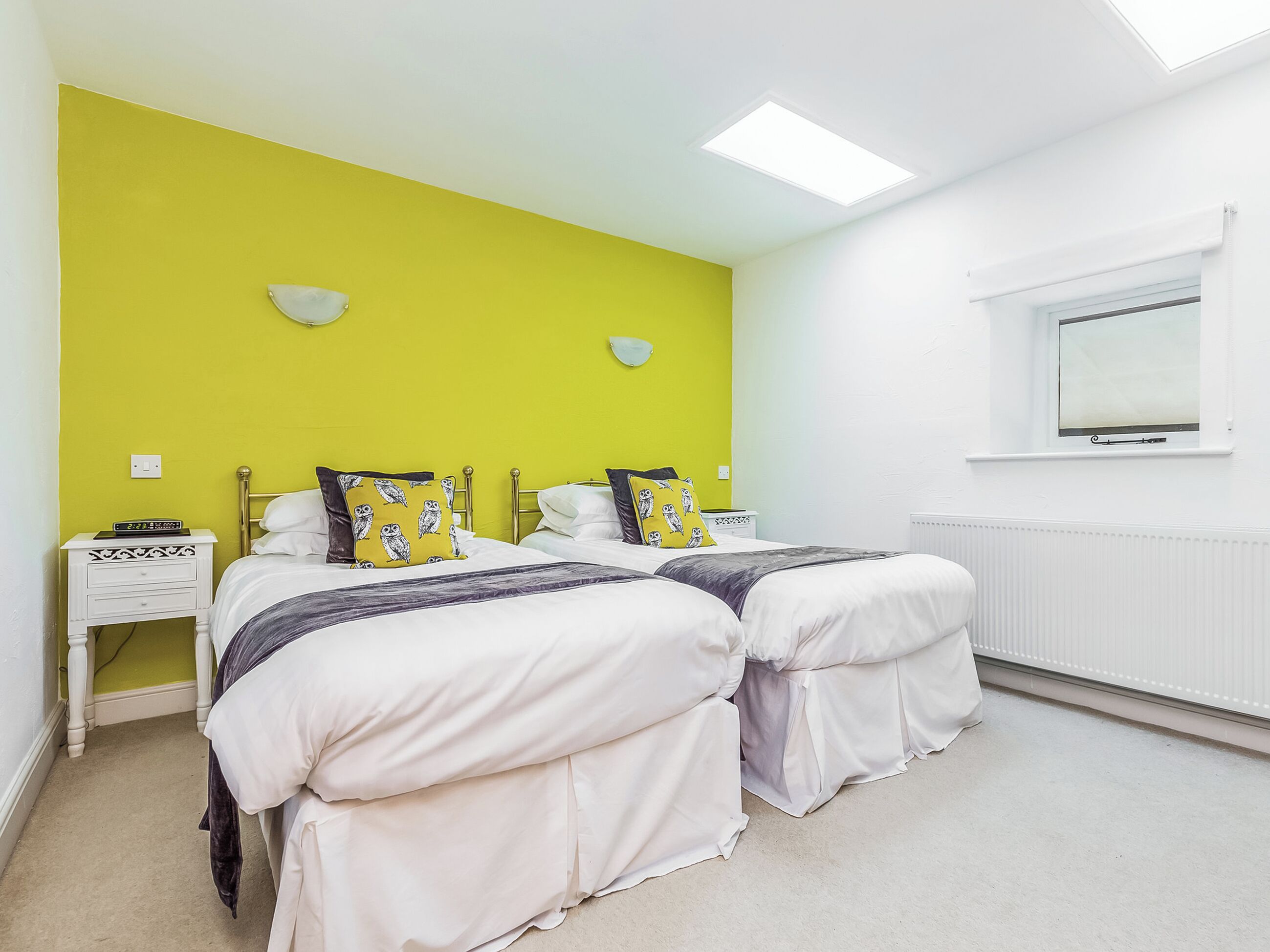 A room with two beds, a white nightstand with a clock, a window with a view, and a radiator.