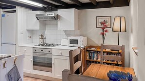 A compact kitchen with white cabinets, a stove, oven, microwave, and a dining area with a wooden table and chairs. - A cute cottage for two, set peacefully with regular red squirrel visitors. (Llaneilian, near Amlwch)