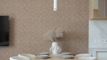 Un coin repas avec une table en bois, des chaises blanches, un vase contenant des fleurs séchées et un téléviseur fixé au mur.