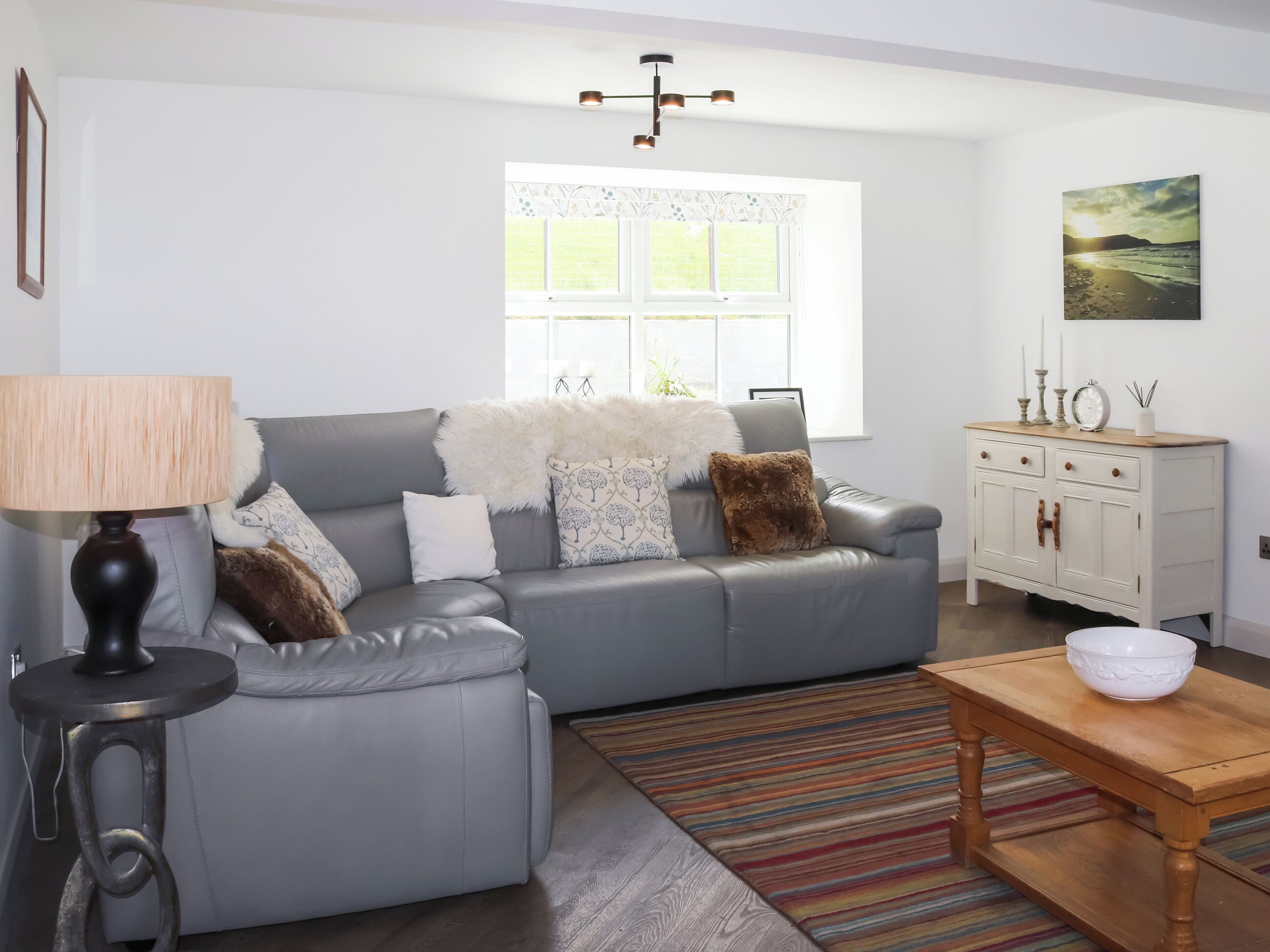 A modern living room with a grey sofa, a wooden side table, a lamp, a patterned rug, and a window with a view.