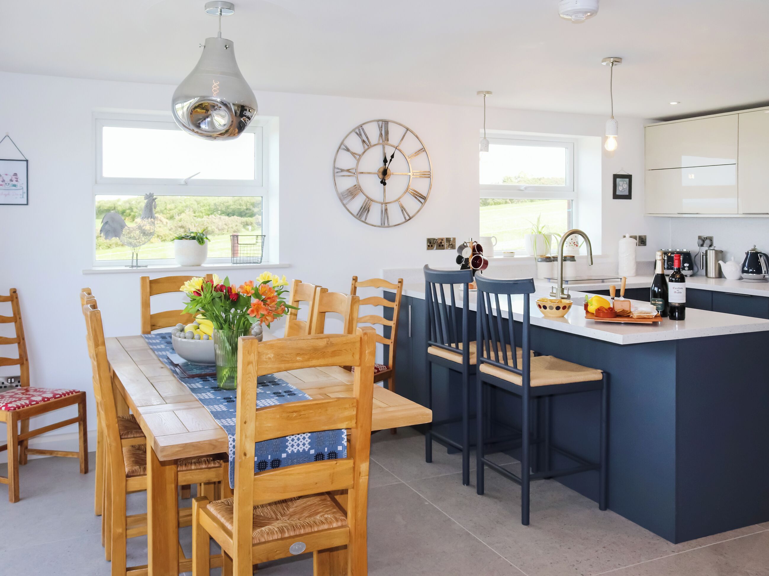 A modern kitchen with a central island, wooden chairs, a dining table with a vase of flowers, and a large wall clock.
