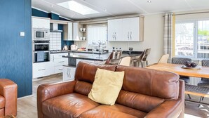A modern kitchen with white cabinets, a dining area with a wooden table, and a brown leather sofa with a pillow. - Serenity at Lakeside - UK51947 (Patrington Haven)