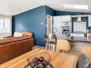 Una sala de estar moderna con un sofá de cuero marrón, una mesa de comedor con un cuenco de fruta y una cocina con gabinetes blancos y una estufa.