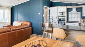 A modern living room with a brown leather sofa, a dining table with a bowl of fruit, and a kitchen area with white cabinets and a stove. - Serenity at Lakeside - UK51947 (Patrington Haven)