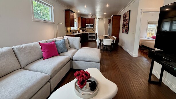 Una sala de estar moderna con un sofá seccional, una mesa de centro con objetos decorativos, una zona de comedor con mesa y sillas, una cocina con electrodomésticos integrados, y un televisor sobre un mueble.