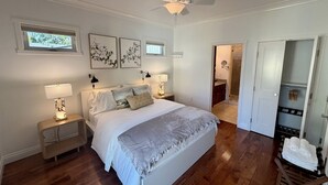 Un dormitorio con cama, mesitas de noche, decoración en la pared, ventilador de techo y una puerta que da a otra habitación.