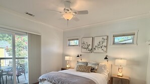 Un dormitorio con una cama, dos mesitas de noche con lámparas, un ventilador de techo y un ventanal corredizo que da a un espacio exterior.