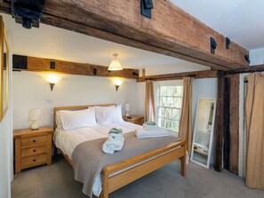 Ein Schlafzimmer mit einem hölzernen Bett, Nachttischen, einem kleinen Tisch und einem Fenster mit Vorhängen, sowie einem Holzbalken an der Decke.