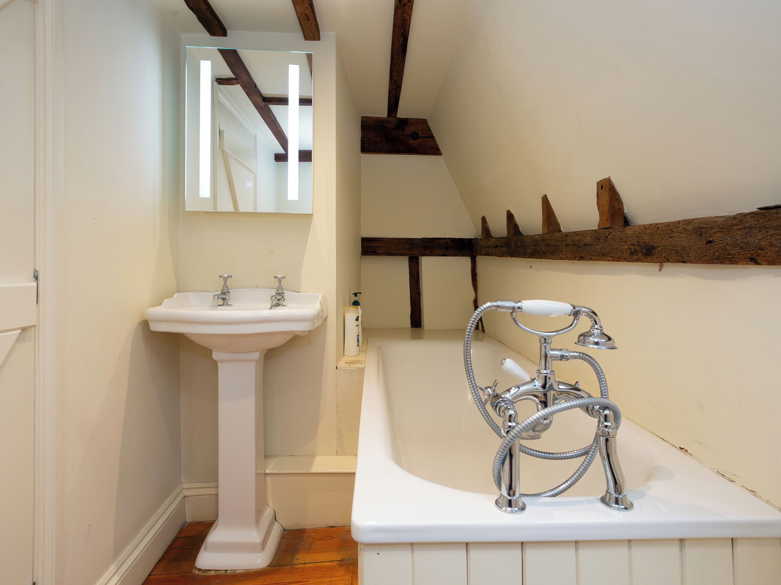 Un baño con lavamanos pedestal, bañera con ducha y vigas de madera en el techo.