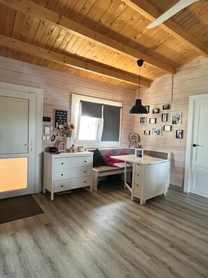 Une pièce avec un plafond en bois, une fenêtre avec des stores, un bureau, un banc et un mur orné de photos encadrées.