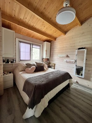 Ein gemütliches Schlafzimmer mit Holzbalkendecke, einem großen Bett mit brauner Decke, einem Fenster mit Jalousien und einem Spiegel mit Beleuchtung.