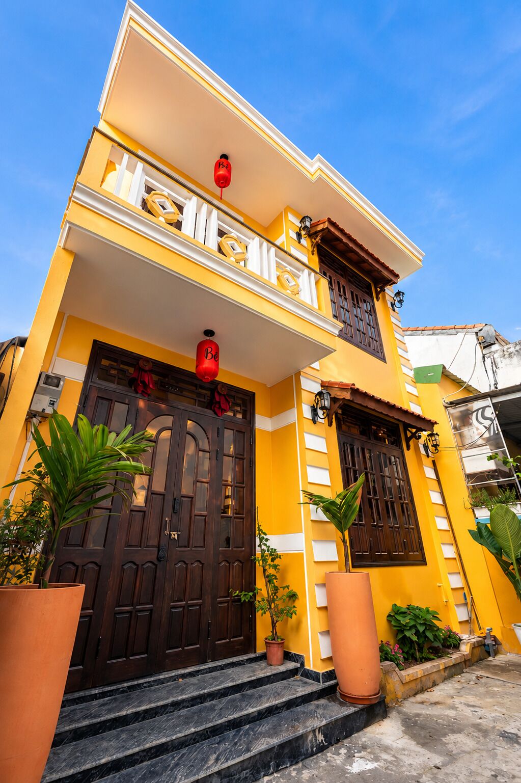 A yellow building with a dark brown door, a balcony, and potted plants.