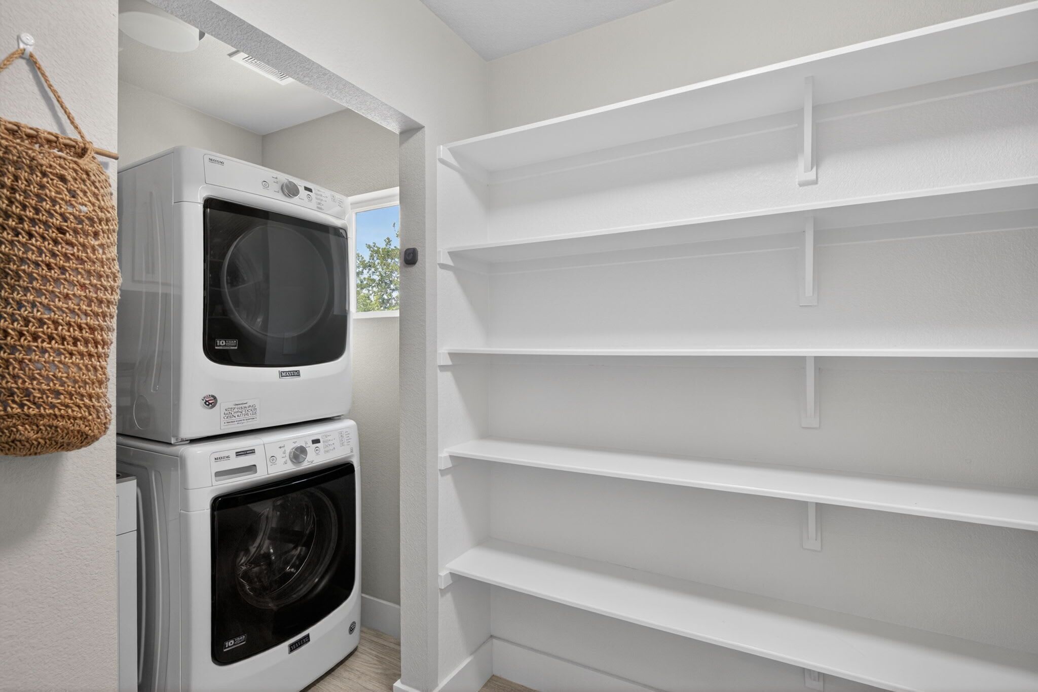 A laundry room with stacked washers and dryers, white shelving, and a hanging basket.