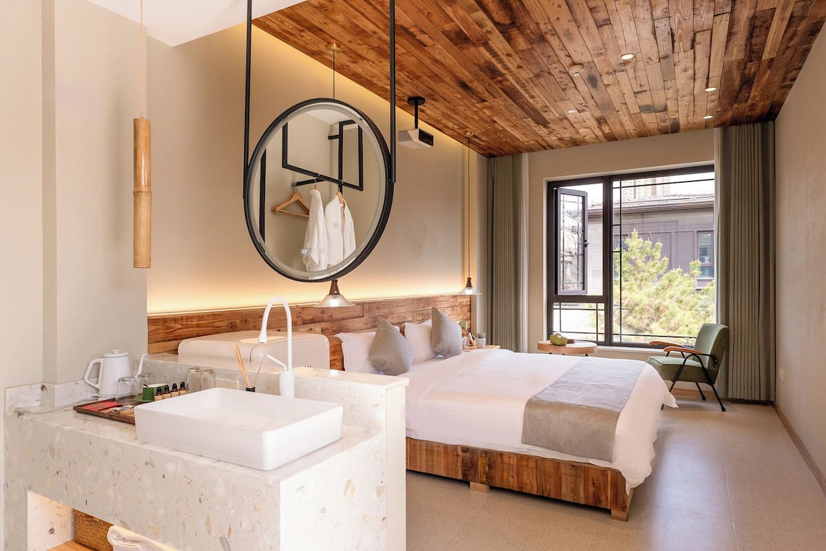 A hotel room with a wooden ceiling, a large round mirror, a bed with a wooden headboard, a bedside table with a white sink, and a view of the outside through a large window.