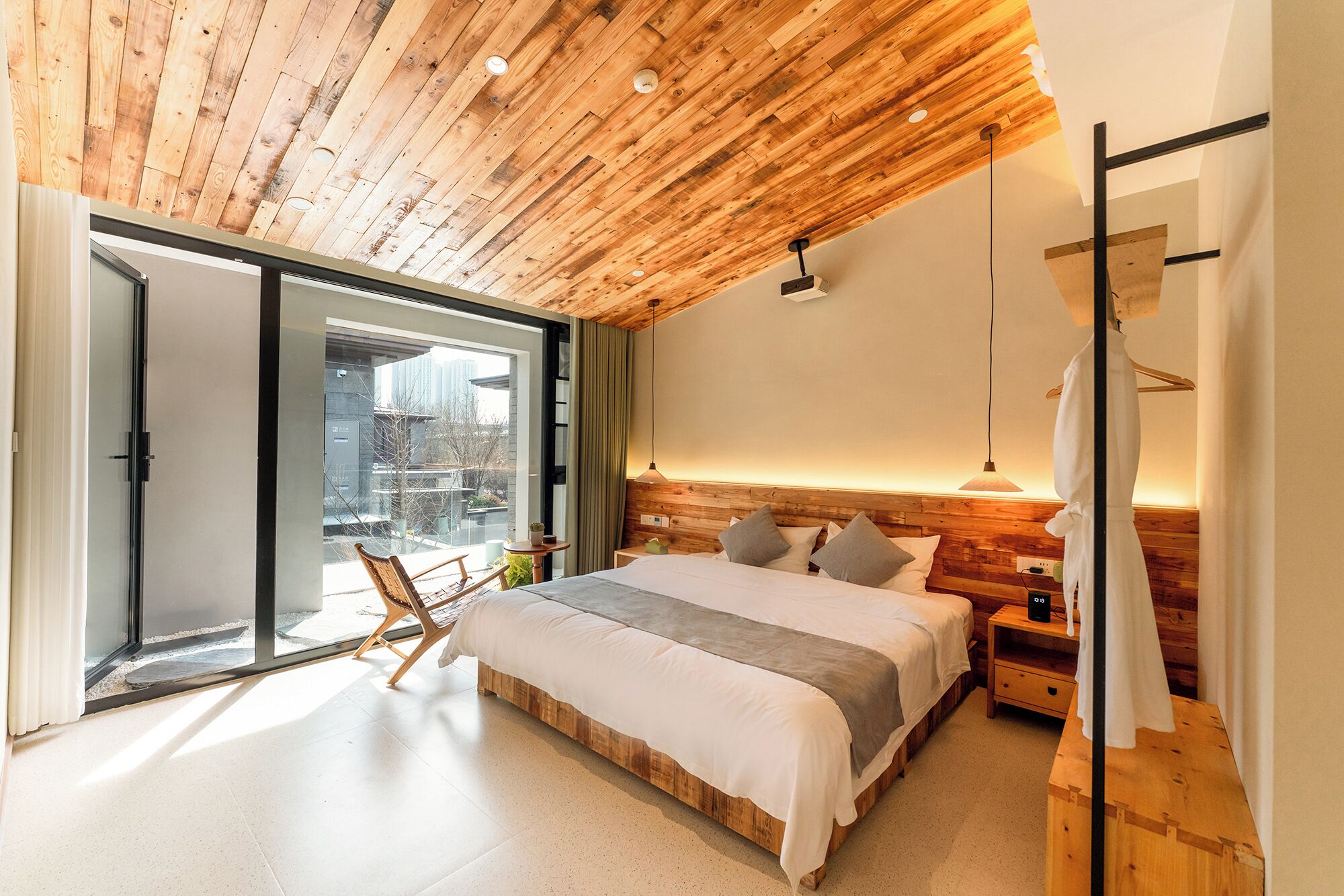 Un dormitorio moderno con techo de madera, una cama grande, una mesita de noche, una silla y vistas a través de un ventanal.