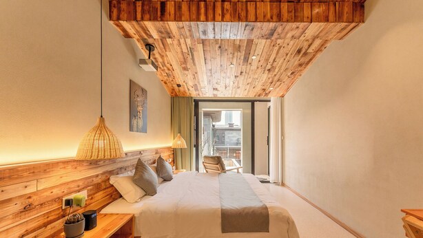 Ein Schlafzimmer mit Holzbalkendecke, einem Bett mit Kissen, einem Nachttisch mit Lampe und Blick durch eine Glastür.