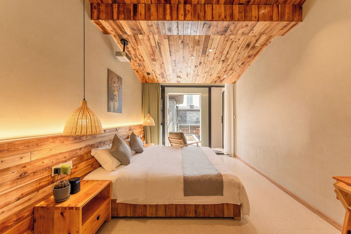 A bedroom with a wooden ceiling, a bed with pillows, a bedside table with a lamp, and a view through a glass door.