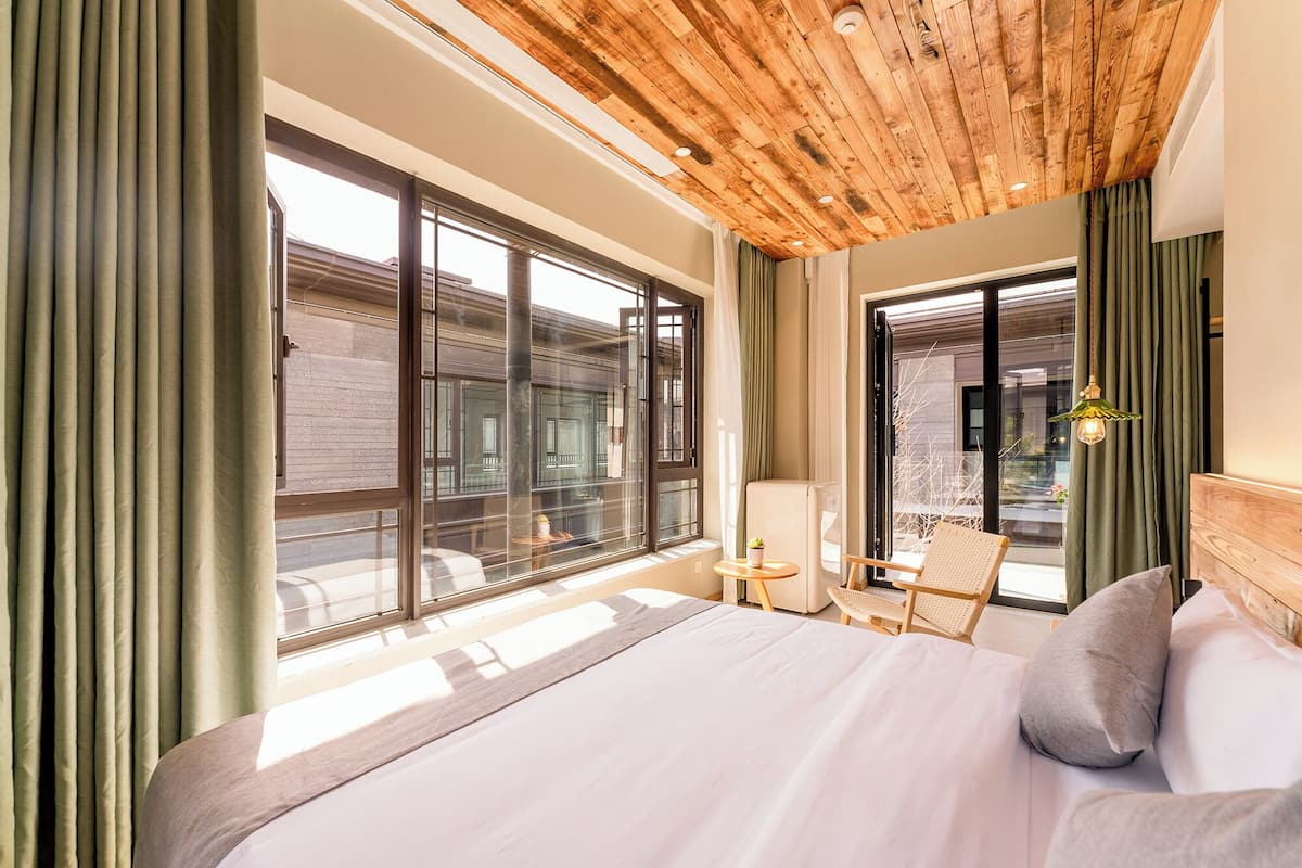 A hotel room with a large bed, a wooden ceiling, and a view of buildings through large windows.